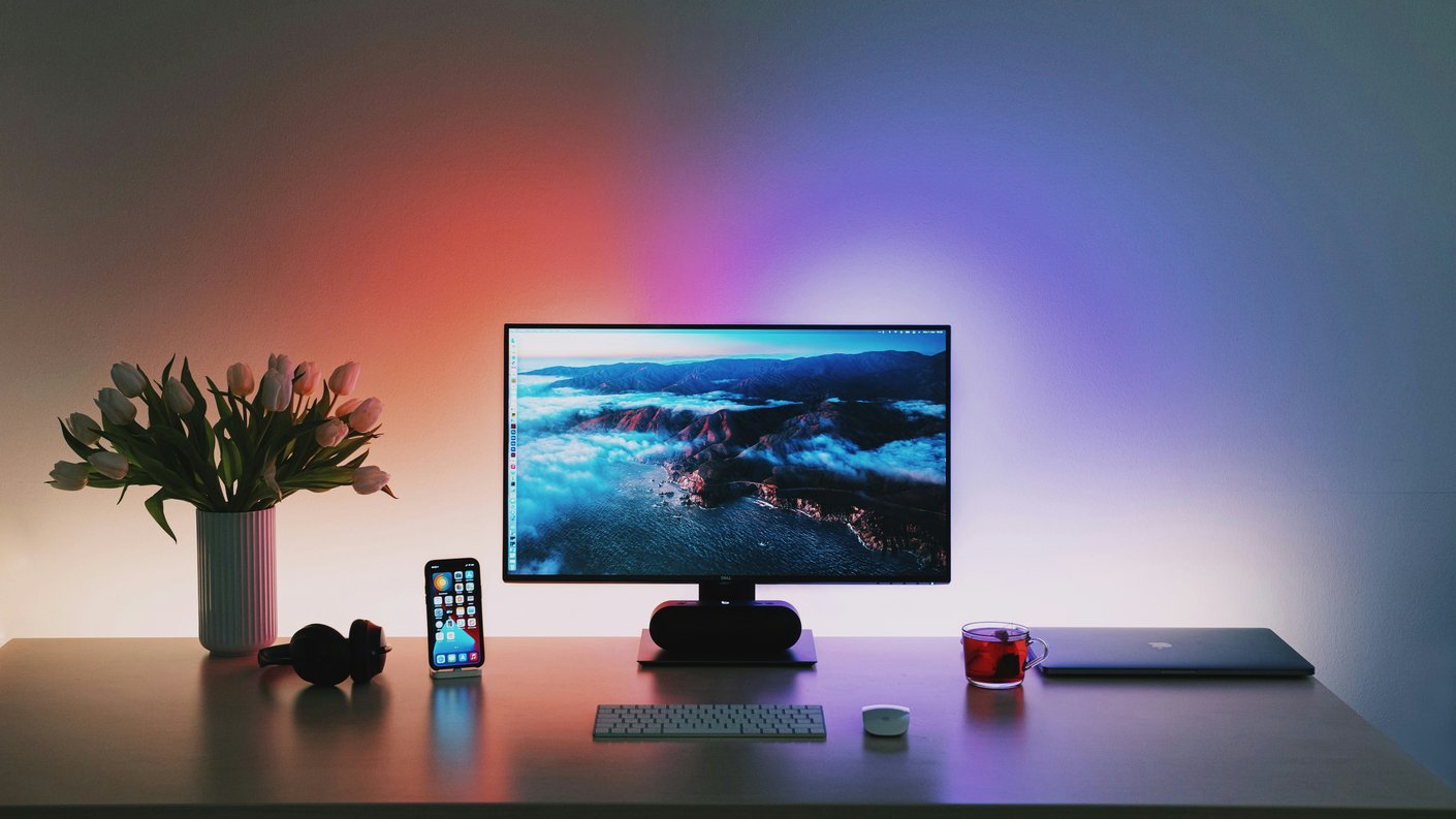 A tidy desk under coloured backlight with a monitor, headphones, a phone and a vase of tulips