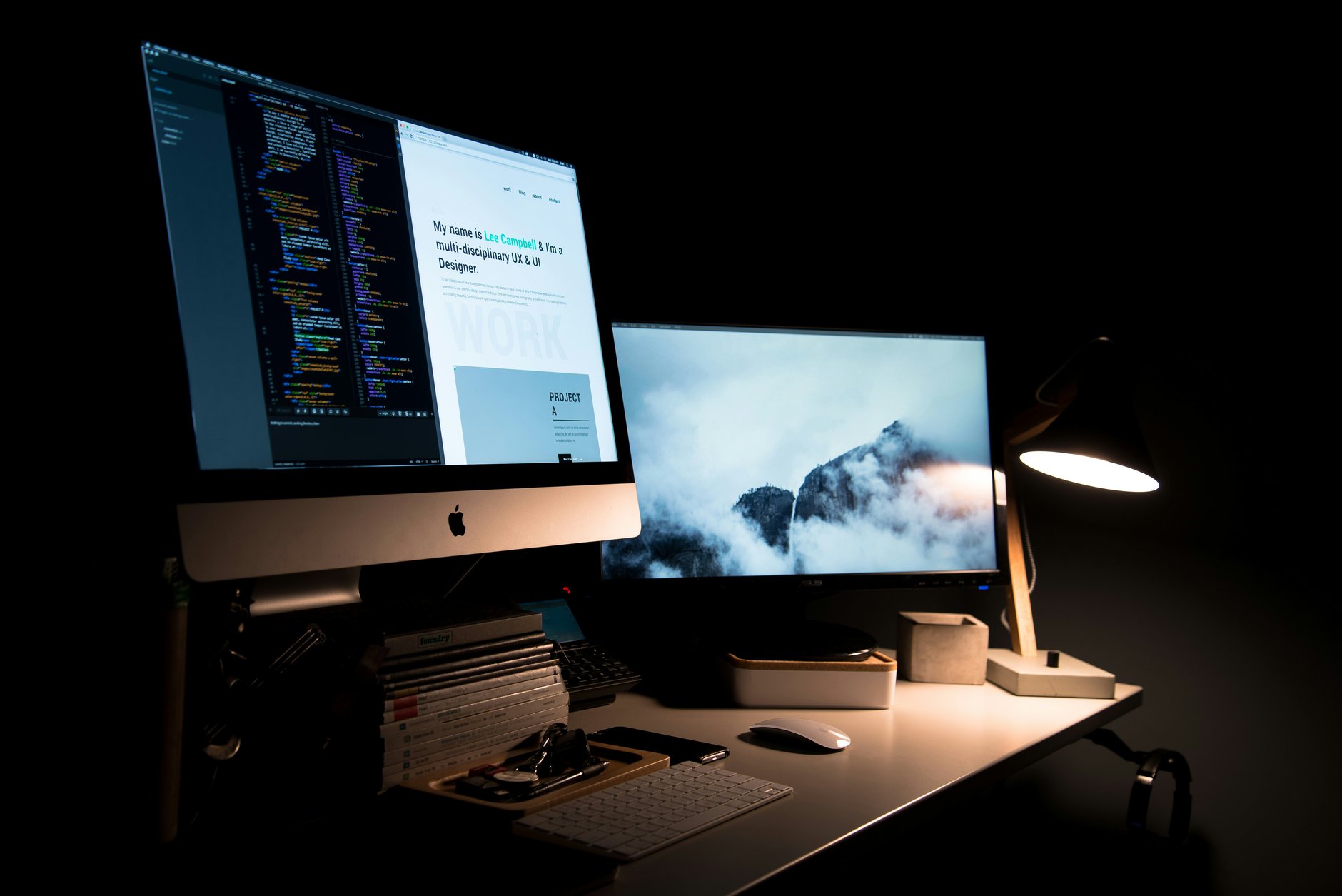A darkened desk at night with two screens showing a design layout and lines of code beside a single lamp